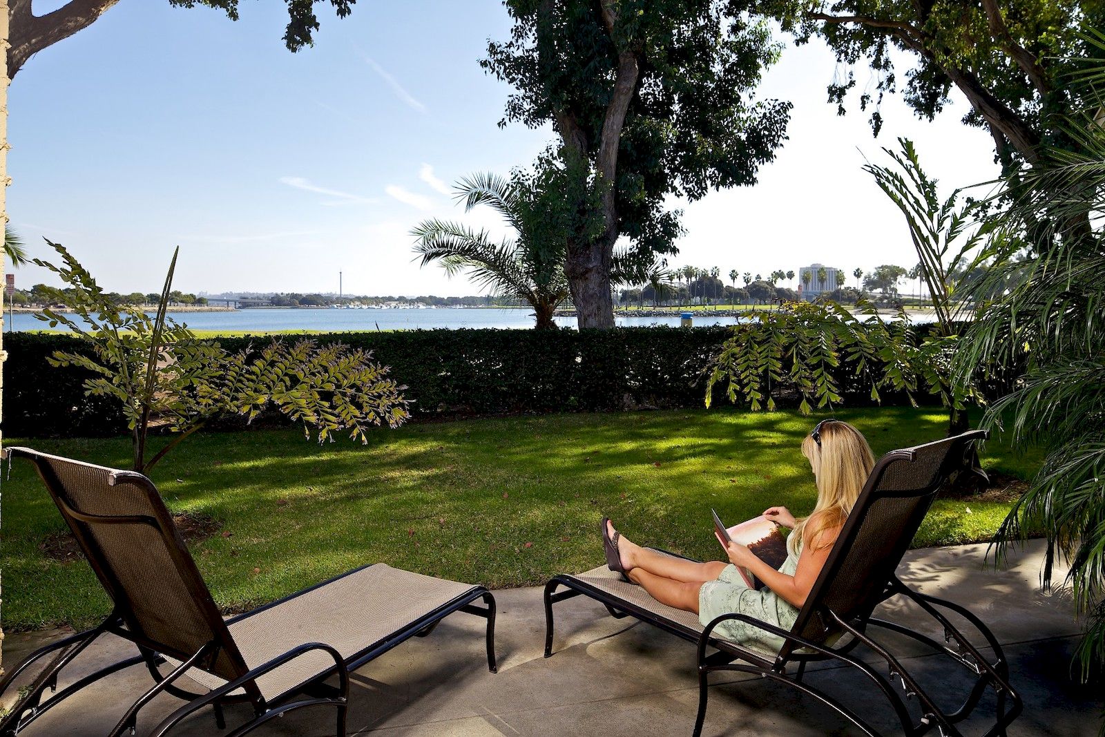 A woman lounging by a lawn, reading a book on a chair near a poolside with trees, water, and distant skyline in the background.