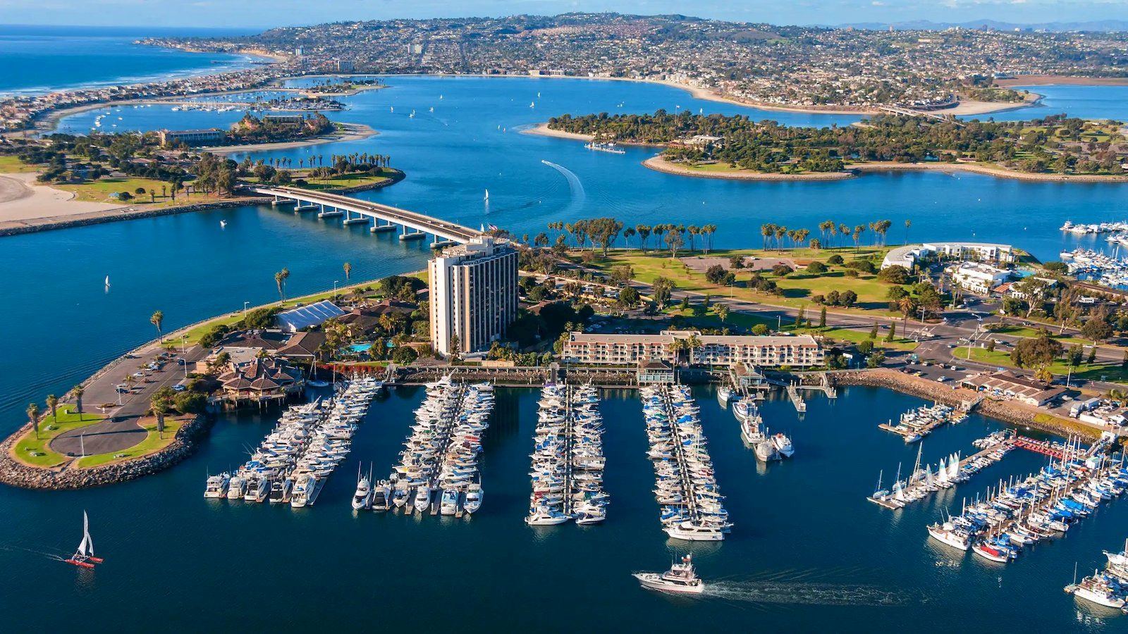 Aerial view of a marina with numerous sailboats docked, a modern building, a long bridge, and blue water in a coastal harbor.
