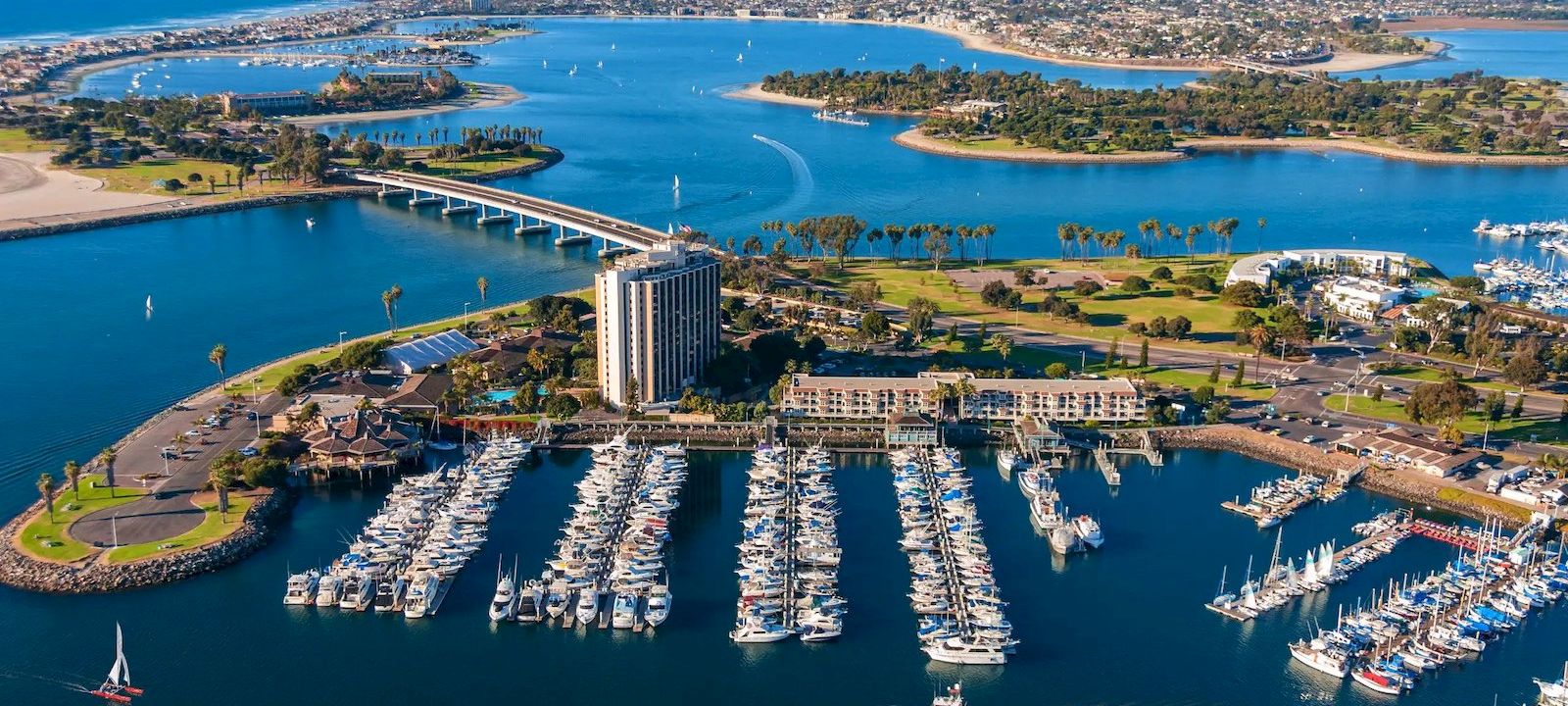 Aerial view of a marina with numerous sailboats docked, a modern building, a long bridge, and blue water in a coastal harbor.