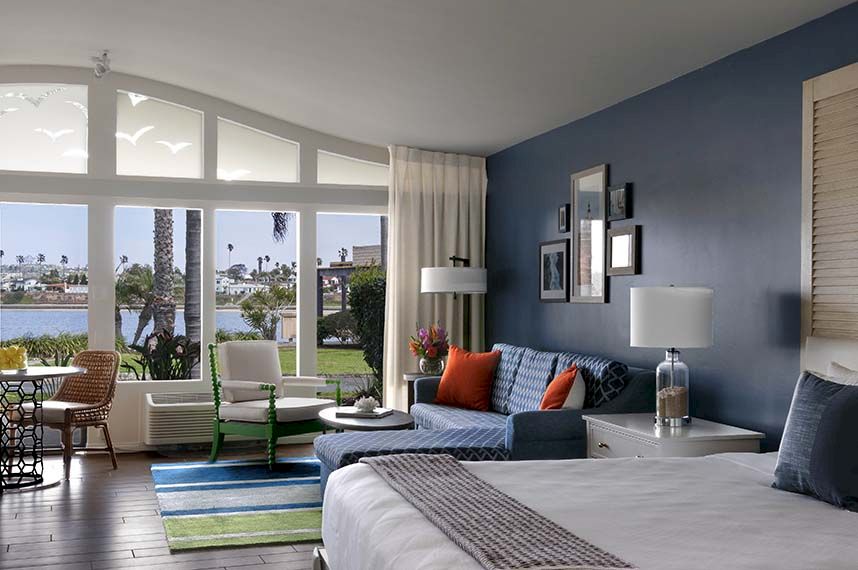 A bright hotel suite with a blue accent wall, large windows opening to a balcony, a bed, a blue sofa with orange pillows, and stylish lamps.
