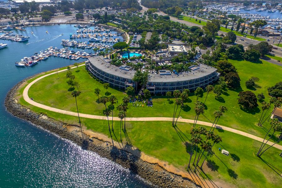 An aerial view of a circular hotel on the coastline, surrounded by green lawns, palm trees, a winding path, and a marina with many boats anchored nearby.
