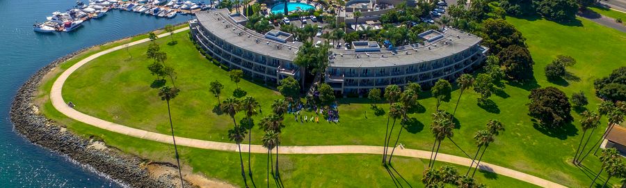 An aerial view of a circular hotel on the coastline, surrounded by green lawns, palm trees, a winding path, and a marina with many boats anchored nearby.