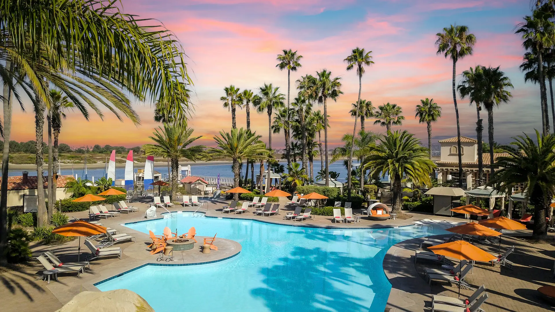A tropical resort pool scene at sunset, with palm trees, lounge chairs, umbrellas, and a calm, blue pool reflecting the sky.