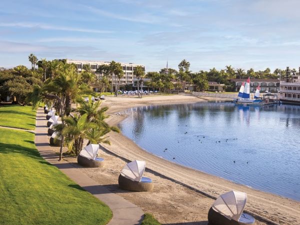 A sunny seaside park with palm trees, green lawns, and a sandy beach along a calm bay; decorative white sail-shaped benches line the shore.