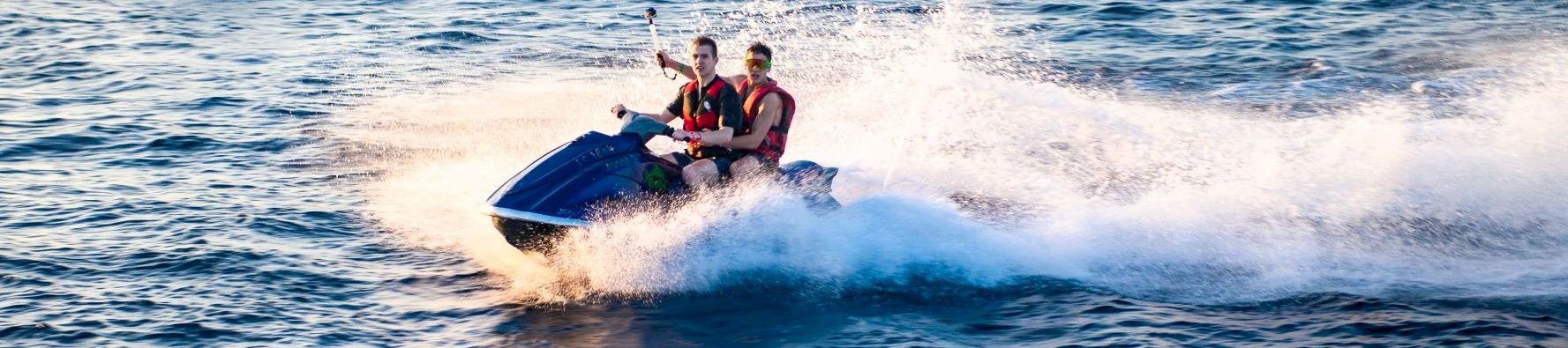 Two people ride a jet ski at high speed on choppy blue water, leaving white spray behind them, with another jet ski in the background.