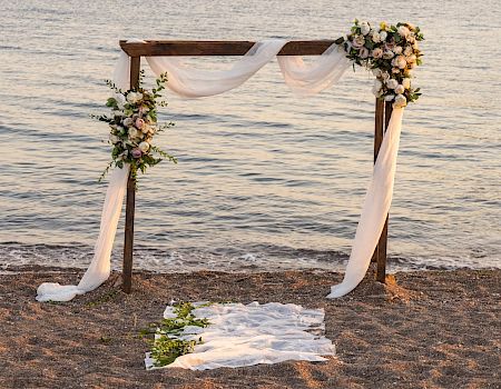 A seaside wedding arch of dark wood draped in white fabric, topped with pale flowers and greenery for a ceremony.