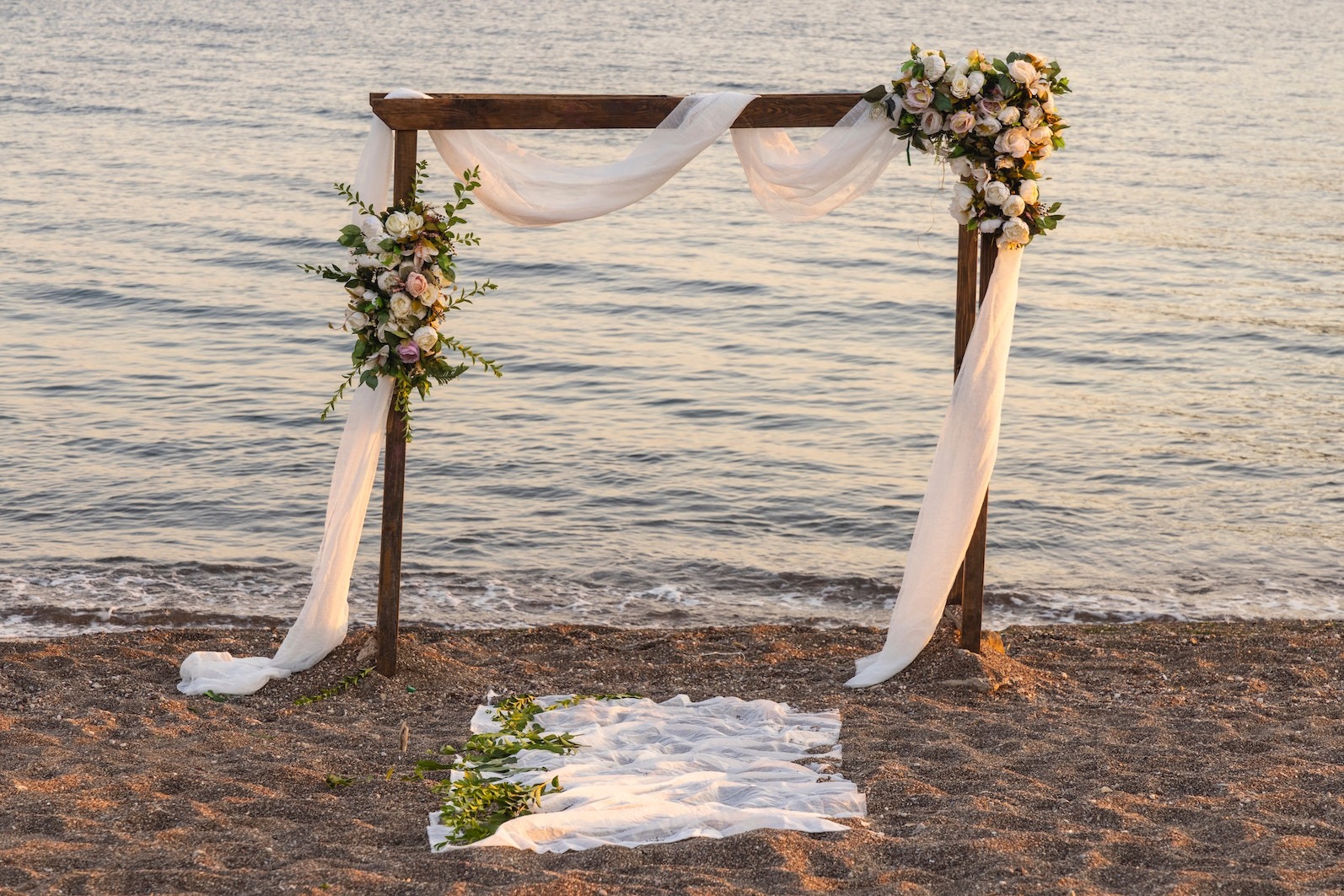 A seaside wedding arch of dark wood draped in white fabric, topped with pale flowers and greenery for a ceremony.