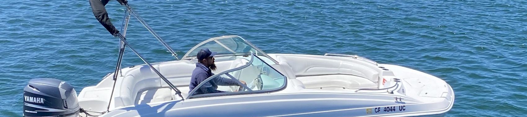 A small white motorboat with a black outboard sits on calm blue-green water; a person wears a cap in the cockpit near a windshield.