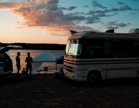 A coastal campsite at sunset with a parked RV, a tent, and a few people near a car, silhouettes against a orange-pink sky ending.