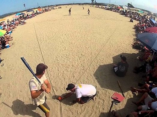 A sand battle at the beach: two people duel with a foam sword while a crowd sits in a wide circle under umbrellas, watching intently.