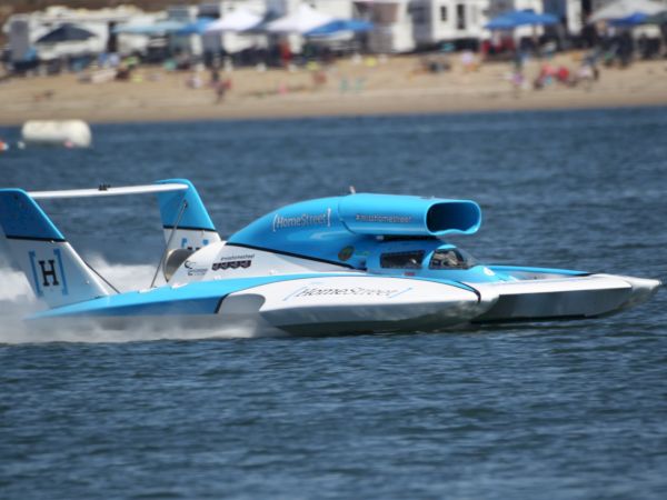 A blue and white hydroplane speedboat glides over water near a beach with people and tents in the background.