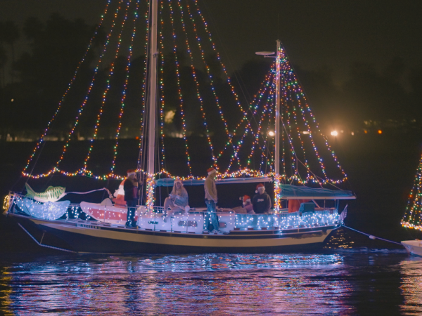 A party sailboat covered in colorful string lights cruises at night, with people on deck and another lit boat nearby.