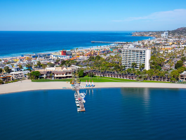 Aerial view of a coastal town with a sandy beach, pier extending into calm blue water, hotels and houses along the shore, and the ocean meeting the horizon.
