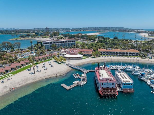 Aerial view of a marina with boats, a sandy beach, palm trees, and a resort complex along the waterfront, bright blue skies, calm turquoise water.