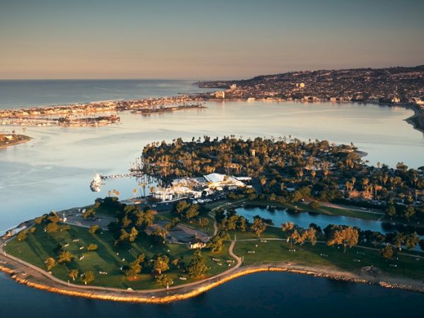 An aerial view of a lush peninsula and marina, with green parks, trees, and a coastline, surrounded by calm blue water and distant cityscape.