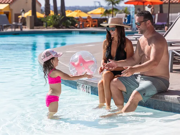 A family by a pool: a little girl in a pink swimsuit splashes water while adults sit on the pool edge, a pink inflatable ball nearby, sunny resort vibe.