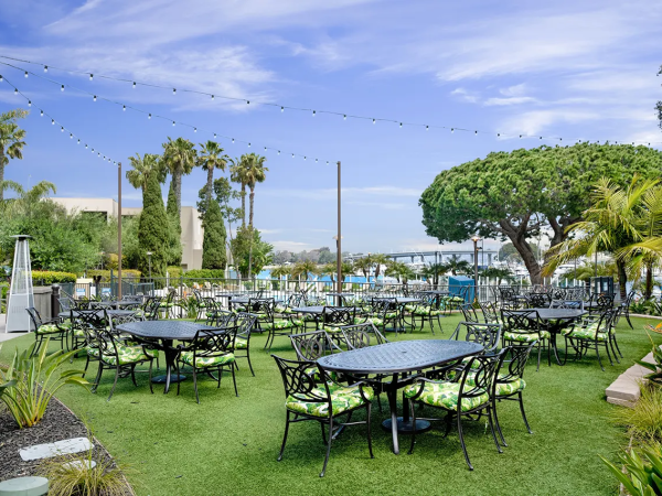 Outdoor cafe with black metal tables and chairs on green artificial turf, string lights, palm trees, and a sunny pool area in the background.