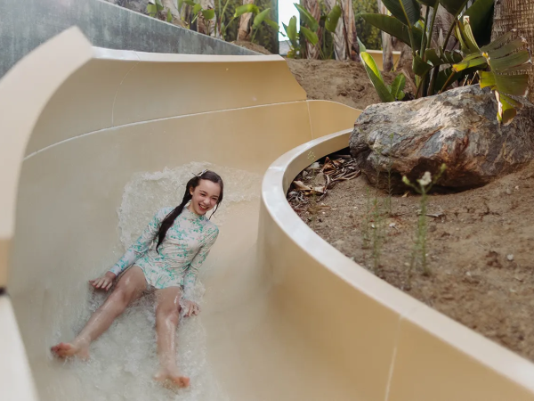 A girl wearing a light blue swimsuit slides down a curved beige waterslide into a splash, surrounded by rocks and tropical plants.