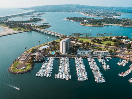 Aerial view of a marina with many white boats, a large building complex, a bridge, and lush green islands in blue water&mdash;vibrant coastal scenery.