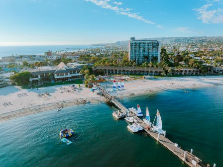 A sunny seaside resort with a long pier, sandy beach, boats, and modern buildings along the coast, under a clear blue sky.