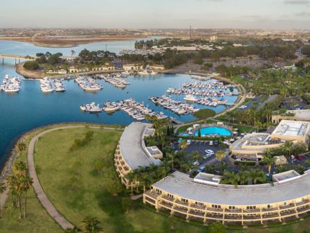 Aerial view of a marina with numerous boats, a curved resort complex, green lawns, and a circular pool area by the water.