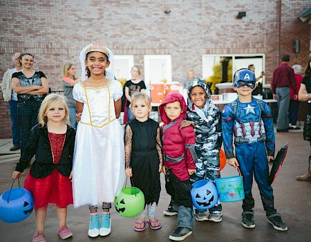 A group of children in Halloween costumes pose together with colorful buckets and jack-o&rsquo;-lantern buckets, smiling outside.