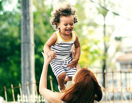 A smiling toddler in a striped outfit is being lifted and held up by an adult outdoors, laughing in a sunny park.