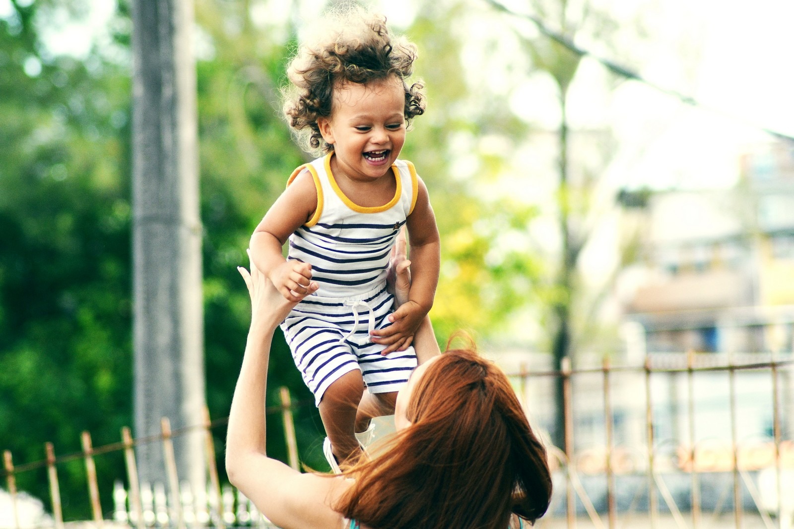 A smiling toddler in a striped outfit is being lifted and held up by an adult outdoors, laughing in a sunny park.