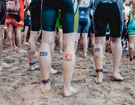 A crowd of runners standing on a sandy beach, wearing numbered ankle bands and athletic shorts, ready for a race.