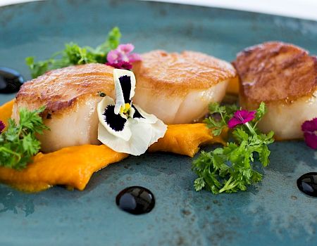 Seared scallops with orange pur&eacute;e, microgreens, edible flowers, and a drizzle of balsamic glaze on a blue plate.