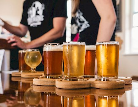 Three pints of beer on a wooden flight board with two bartenders in the background, indoors, casual bar scene.