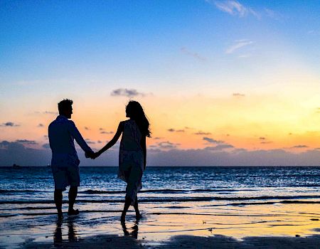 Two people holding hands and walking along the beach at sunset, silhouettes against a colorful sky.