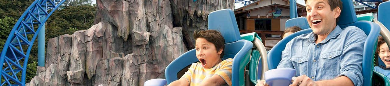 People ride a blue roller coaster at a theme park, screaming with excitement as a man and a boy hold on tight aboard the ride.