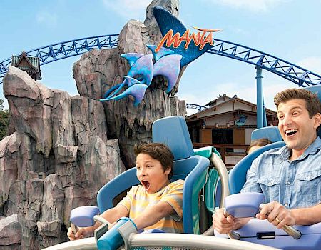 People ride a blue roller coaster at a theme park, screaming with excitement as a man and a boy hold on tight aboard the ride.