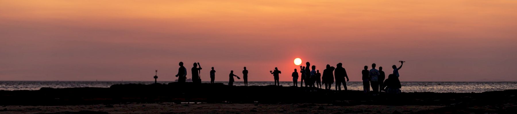 A group of people on a beach at sunset, silhouettes against an orange sky, some standing, some taking photos, horizon over calm water.