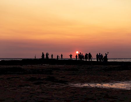 A group of people on a beach at sunset, silhouettes against an orange sky, some standing, some taking photos, horizon over calm water.