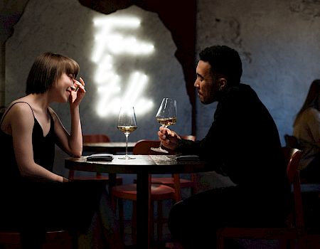 Two people sit per table in a dim, moody restaurant; a man and woman share a wine moment while others blur in the background, lights glow softly.