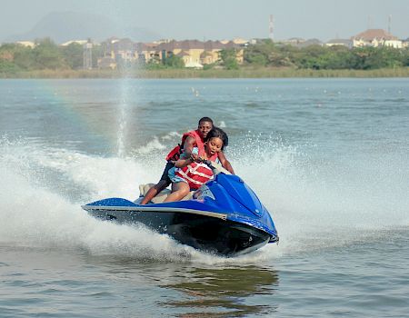 A couple rides a blue jet ski fast across a lake, creating a spray as they hold on tightly and smile, with a distant shoreline in view.