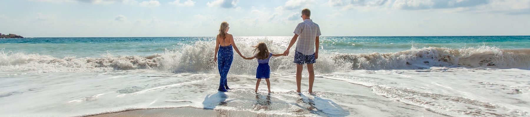 A family of three stands at the shore as waves roll in, holding hands on a sunny beach with blue skies.