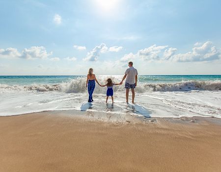 A family of three stands at the shore as waves roll in, holding hands on a sunny beach with blue skies.