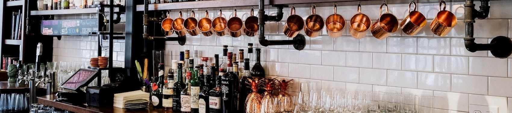A well-stocked bar with bottles on shelves, copper mugs, and row of hanging glasses behind a polished wooden counter, ready for service.
