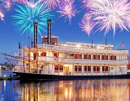 A large, multi-deck riverboat on calm water with colorful fireworks overhead, reflecting in the water at dusk.