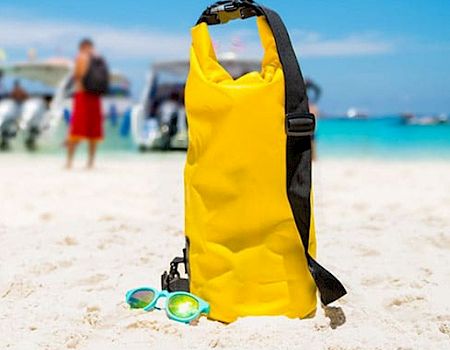 A yellow dry bag and goggles on white sand at the beach, with boats and people in the turquoise water in the background.