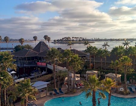 A tropical resort pool area with palm trees, lounge chairs, a thatched-roof bar, and a sandy beach beyond, under a partly cloudy sky.