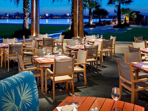 An upscale outdoor restaurant at dusk with wooden tables, cushioned chairs, glassware, and a view of palm trees and a lake or ocean backdrop.