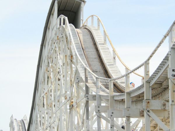 White roller-coaster track climbs into a tower with a dark tunnel-like section and curved support beams against a blue sky.