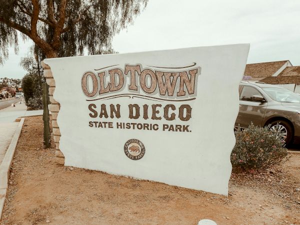 A sign for &ldquo;Old Town San Diego State Historic Park&rdquo; stands beside a sidewalk and parked cars.