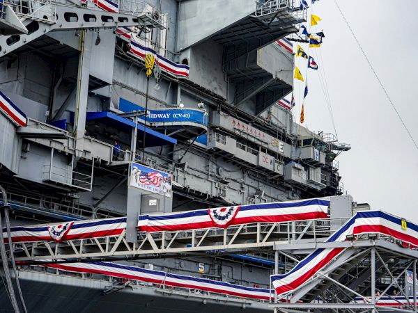 A Navy aircraft carrier deck packed with flags and the blue sign &ldquo;MIDWAY (CV-41),&rdquo; with large cranes and gray steel.