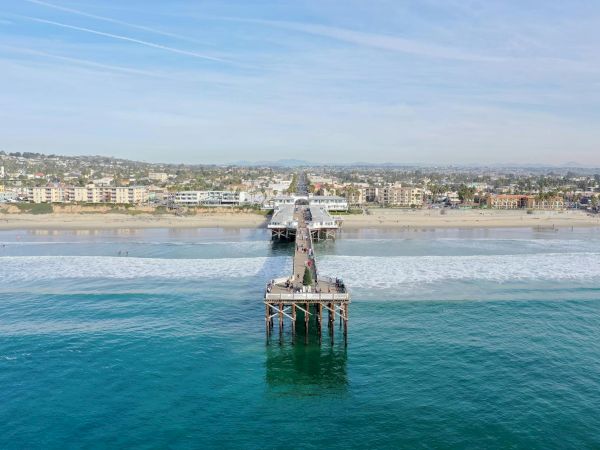 A long wooden pier extends into the ocean off a sandy beach, with buildings and swimmers along the coastline under a blue sky.