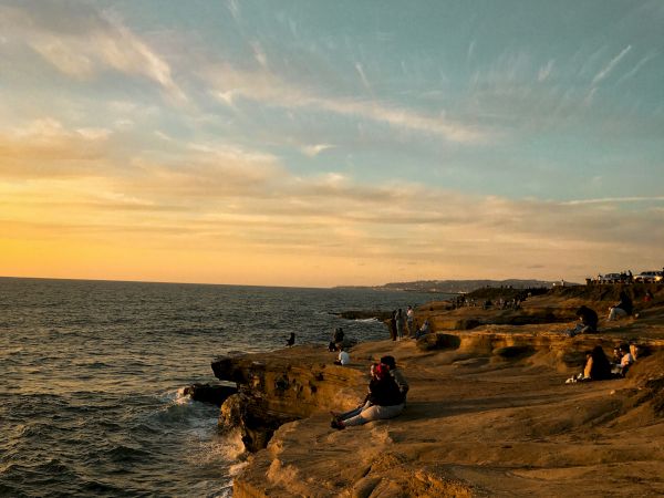 A golden sunset over the ocean cliffs, with people relaxing and watching the waves crash below.